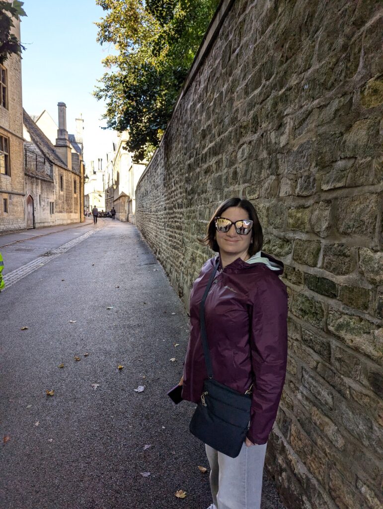 Three quarters profile of Lisa posing for the camera wearing a plum colored waterproof jacket, light jeans, sunglasses, and a crossbody bag. She's standing beside a stone wall on a narrow historic street in Oxford lined with old stone buildings and trees.